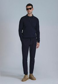 Navyblaues, geripptes Langarm-Polo-Shirt, kombiniert mit passenden, taillierten Hosen und hellbraunen Wildlederschuhen. Die Accessoires umfassen dunkle Sonnenbrillen.