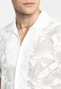 Witte korte mouwen shirt met een geometrisch open breipatroon met verschillende texturen en subtiele beige accenten. Knopsluiting bij de kraag.