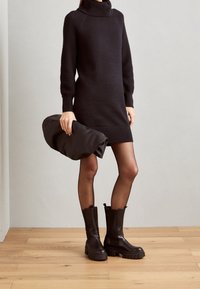 Robe pull noire avec manches texturées, col haut et silhouette cintrée. Assortie à des bottines marron à semelles épaisses. Tenant un sac noir.