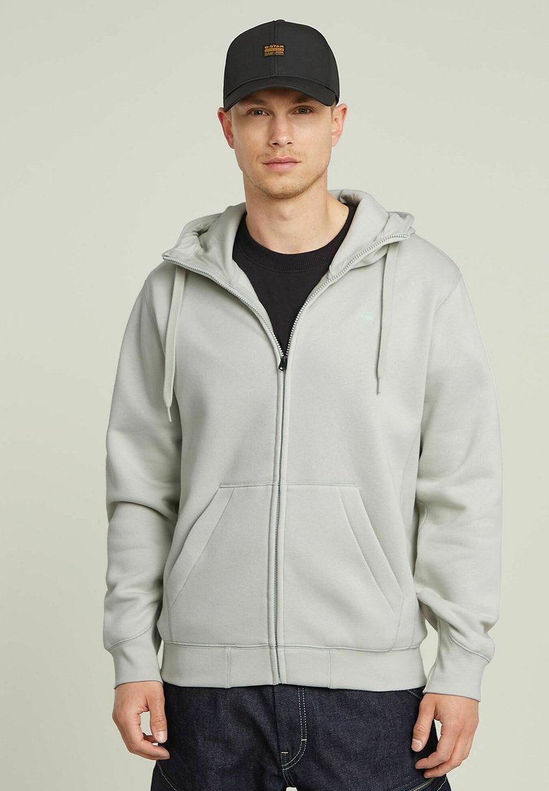 Un sweat à capuche zippé gris clair avec des cordons de serrage, deux poches avant et des poignets côtelés, associé à une casquette noire arborant un logo.