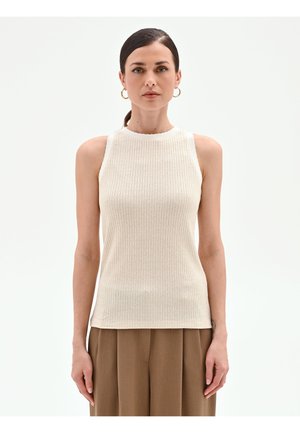 Femme aux cheveux bruns attachés en queue de cheval, portant des boucles d'oreilles créoles en or, un haut sans manches beige en maille côtelée, et un pantalon plissé marron, sur fond blanc.