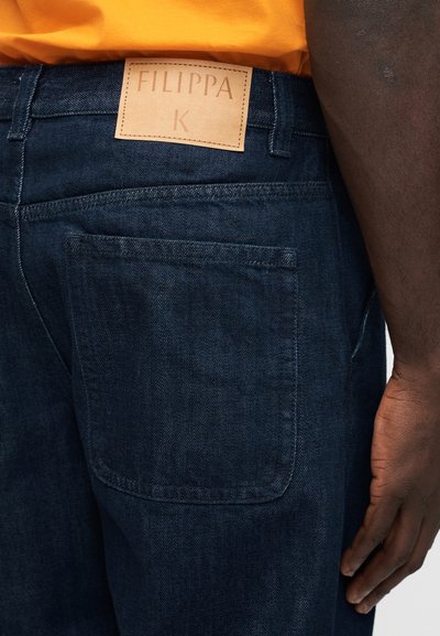 Πίσω τσέπη από σκούρο μπλε τζιν και ταμπόν από ταμπά δέρμα με την επιγραφή «FILIPPA K», φορεμένα με φωτεινή πορτοκαλί μπλούζα σε άτομο με σκουρόχρωμο δέρμα.