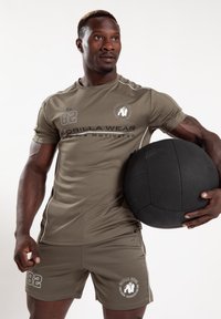 Sportoutfit bestaat uit een aansluitend olijfgroen shirt en shorts, beide met witte accenten en logo's, vastgehouden met een zwarte gewichtenbal.