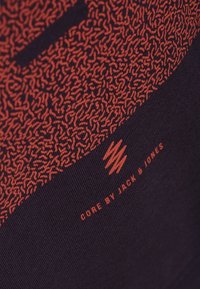 Tela negra con patrones texturizados en rojo y la marca "CORE by JACK & JONES" en una fuente roja en contraste en la parte inferior.