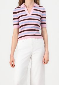 Polo s krátkým rukávem pletené v růžových, modrých a hnědých pruzích, s výstřihem do V a žebrovanou strukturou. V kombinaci s bílými kalhotami s širokými nohavicemi.