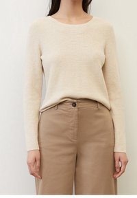 Beige ribattisetakki, jossa on pyöreä kaula-aukko ja pitkät hihat, yhdistettynä korkeavyötäröisiin vaaleanruskeisiin puuvillahousuihin, joissa on edessä nappisulku.