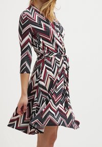 F Wrap klänning med en chevronmönster i vinröd, svart och kräm, med trekvartsärmar och ett knytbälte, tillverkad av ett mjukt tyg.