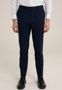 Jūras spēku stila pusbikses ar šauru piegriezumu, taisnu priekšpusi un gludu audumu. Ar vienu pogu aizdari un jostas cilpām. Redzami melni uzvalka apavi.