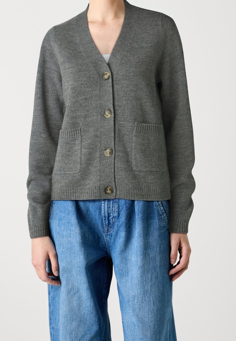 Graue Strickjacke aus strukturiertem Strickstoff, mit V-Ausschnitt, vorderen Taschen und fünf Knöpfen. Kombiniert mit lockeren blauen Jeans.