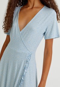 Femme portant une robe portefeuille bleu clair avec un petit motif floral blanc, manches courtes et un détail volanté le long de la couture de la taille.