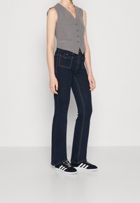 Person som bär en grå ärmlös väst med knappar, mörkblå jeans med framfickor och svarta sneakers med vita ränder.