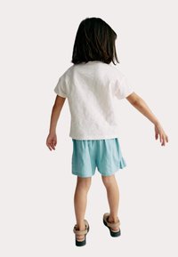 Vit bomull T-shirt, ljusblå shorts med texturerad väv, och svarta sandaler med beiga remmar, sedd bakifrån.