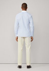 Uomo di spalle che indossa una camicia a righe azzurre chiare, pantaloni color crema e scarpe marroni scure davanti a uno sfondo semplice.