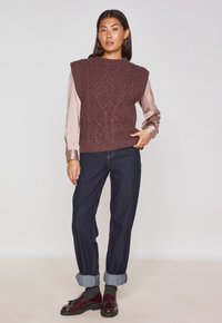 Gilet in magenta a maglia con un motivo a diamante, sovrapposto a una blusa rosa chiaro, abbinato a jeans in denim scuro e mocassini bordeaux.