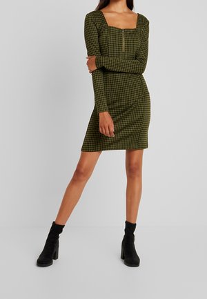 Robe à carreaux verts avec un décolleté carré, manches longues et fermeture éclair à l'avant. Bottines noires à talons complètent le look.