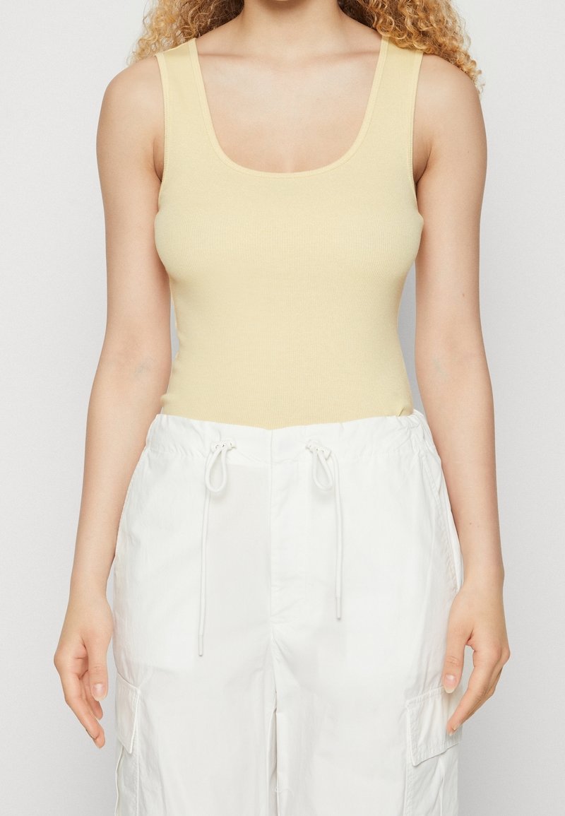 Femme portant un haut sans manches jaune pâle et un pantalon cargo blanc à cordon, montrée des épaules jusqu'aux hanches sur un fond uni.