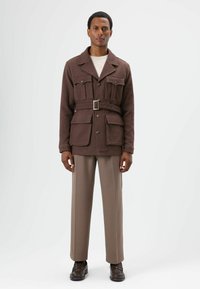 Braune Jacke mit Gürtel und knöpfbaren Taschen, getragen über einem leichten Pullover und beigen Hosen. Glatte Textur, strukturierte Form und maßgeschneiderte Passform.