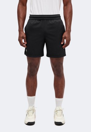 Hombre que lleva pantalones cortos deportivos negros con cintura elástica, calcetines blancos y zapatillas blancas, de pie frente a un fondo claro y liso.