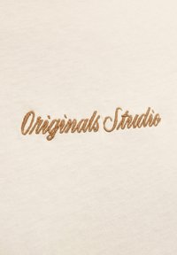 Tissu couleur crème avec du texte brodé "Originals Studio" en cursif marron, présentant une texture douce et lisse.