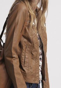 Brązowa kurtka z sztucznej skóry o dopasowanym kroju, z zamkiem błyskawicznym z przodu i dwoma bocznymi kieszeniami. Nosi ją z wzorzystą bluzką, dopełnioną brązową torbą.