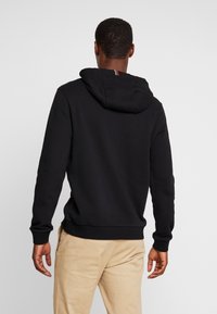 Sudadera negra hecha de una mezcla suave de algodón, con un bolsillo tipo canguro y una capucha con cordón. Destacan las rayas rojas y blancas en el borde de la capucha.