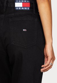 Jeans de mezclilla negra con un bolsillo trasero que lleva una pequeña etiqueta de logo y una etiqueta en la cinturilla que dice "TOMMY JEANS" en rojo, blanco y azul.