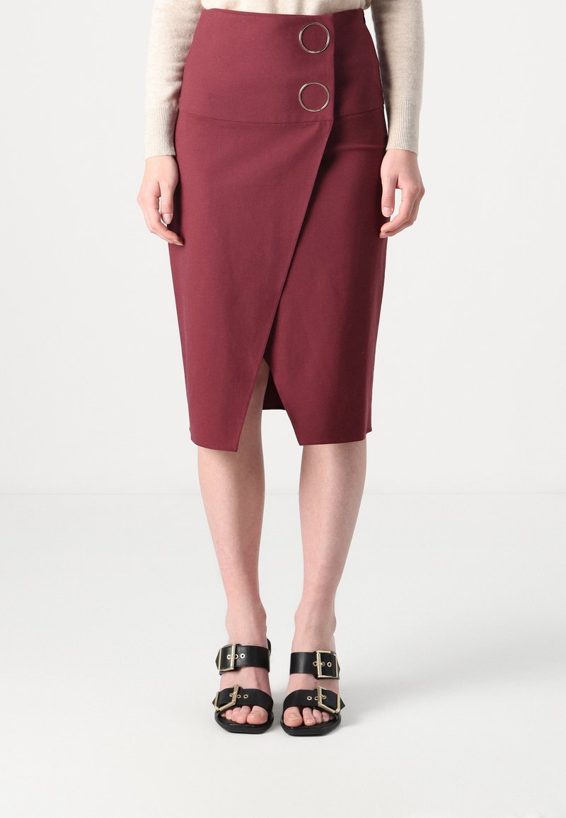Jupe crayon bordeaux avec un ourlet en biais, dotée de deux boutons ronds en métal à la taille. Associée à des sandales noires avec des boucles.