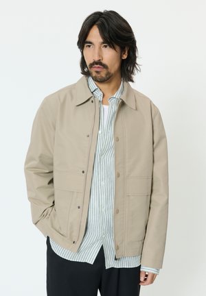 Homme aux cheveux mi-longs noirs portant une veste beige par-dessus une chemise rayée boutonnée et un pantalon noir, regardant vers la gauche.