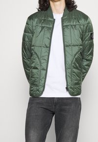 Lätt, vadderad grön bomberjacka med dragkedja, sidofickor och ribbad krage. Bärs över en vit T-shirt och mörka jeans.