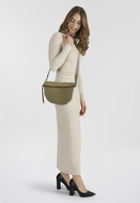 Donna in un vestito lungo beige a maniche lunghe e tacchi neri, che porta una borsa a tracolla verde oliva, in piedi su uno sfondo chiaro e semplice.