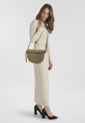 Donna in un vestito lungo beige a maniche lunghe e tacchi neri, che porta una borsa a tracolla verde oliva, in piedi su uno sfondo chiaro e semplice.