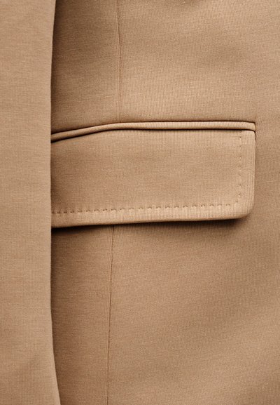 Κοντινό πλάνο μιας τσέπης μπουφάν από ταμπά ύφασμα με καπάκι και εμφανείς ραφές στο υφή υλικό.