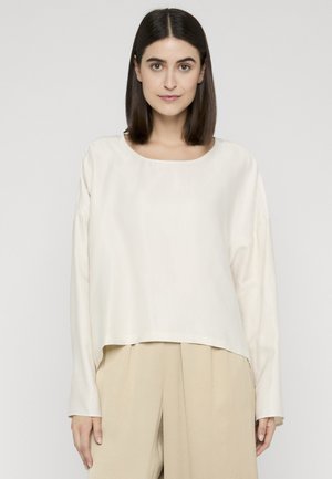 Frau mit glatten, dunklen Haaren trägt ein locker sitzendes cremefarbenes Langarmshirt und beige weite Hosen, steht vor einem schlichten hellen Hintergrund.