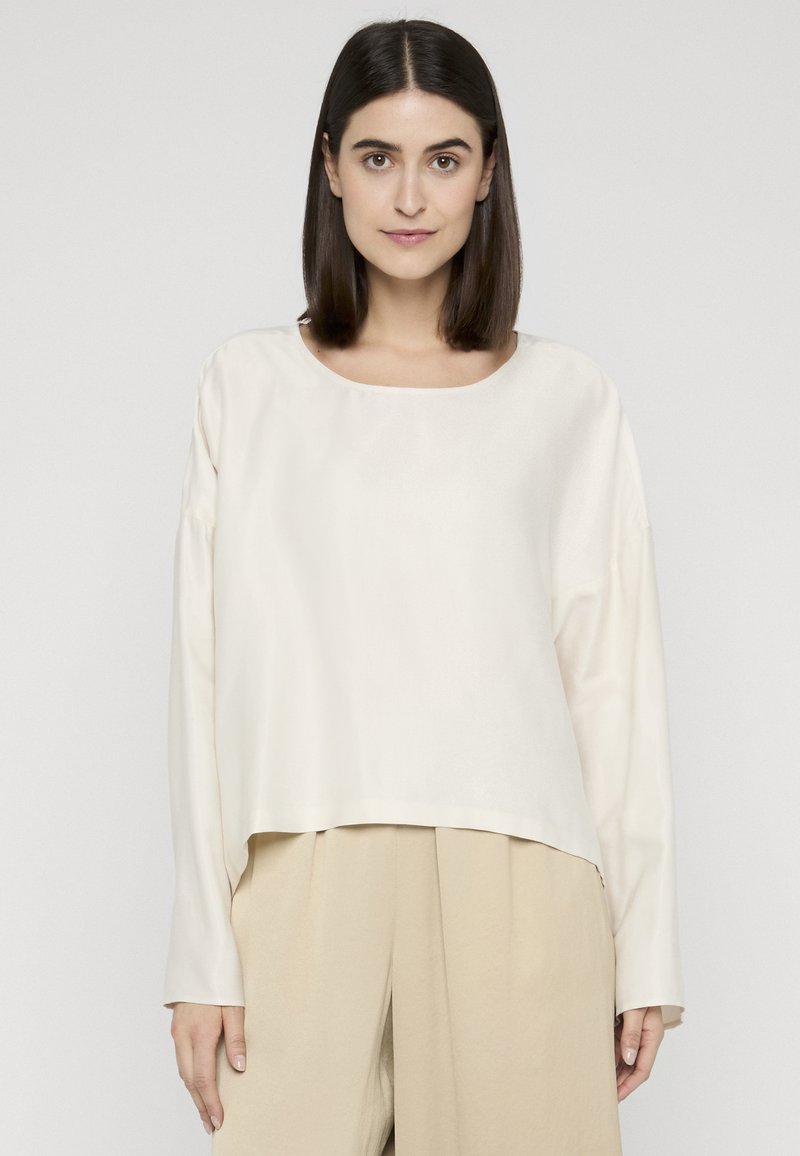Femme aux cheveux raides et foncés, portant un haut ample à manches longues crème et un pantalon beige à jambes larges, debout devant un fond clair uni.