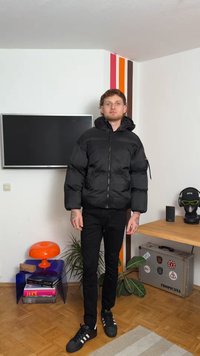 Giacca a vento nera con chiusura a zip e cappuccio, caratterizzata da una texture lucida e polsini elasticizzati, indossata sopra pantaloni neri e scarpe.