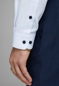 Närbild av en hand som bär en vit, texturerad skjortmudd med två svarta knappar, ett guldarmband och mörkblå byxor.