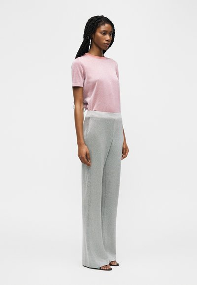 Femme aux cheveux tressés portant un haut à manches courtes rose et un pantalon argenté large à taille haute, debout devant un fond uni.