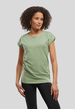 Femme aux cheveux bouclés porte un t-shirt vert à manches courtes avec un petit dessin de main et de cœur, associé à des leggings noirs, debout devant un fond gris.