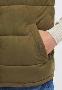 Gilet rembourré vert olive avec une poche latérale à fermeture à bouton. Tissu texturé avec un design matelassé, porté par-dessus un pull gris clair.