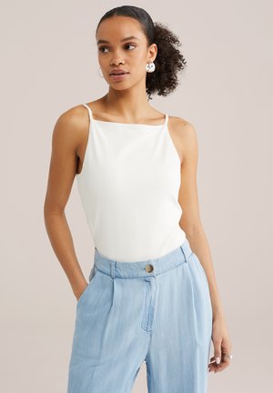 Göndör hajú nő fehér ujjatlan felsőben és világoskék, magasderekú nadrágban, egyik kezét zsebében tartva áll.