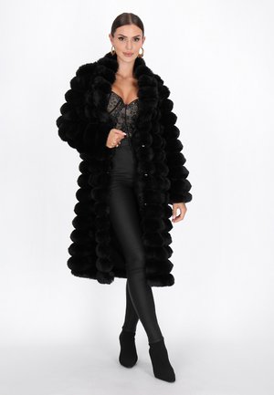 Sort faux fur frakke med rundede puffer, lange ærmer og knaplukning, båret over en sort blonde-top og tætsiddende sorte bukser.
