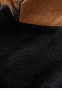 Svart ulltyg med en mjuk textur, med en krage av brunt filt. Designen är minimalistisk utan synlig hårdvara eller mönster.