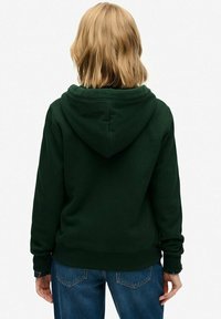 Persona con cabello rubio hasta los hombros, vistiendo una sudadera verde oscuro y jeans azules, vista desde atrás contra un fondo liso.
