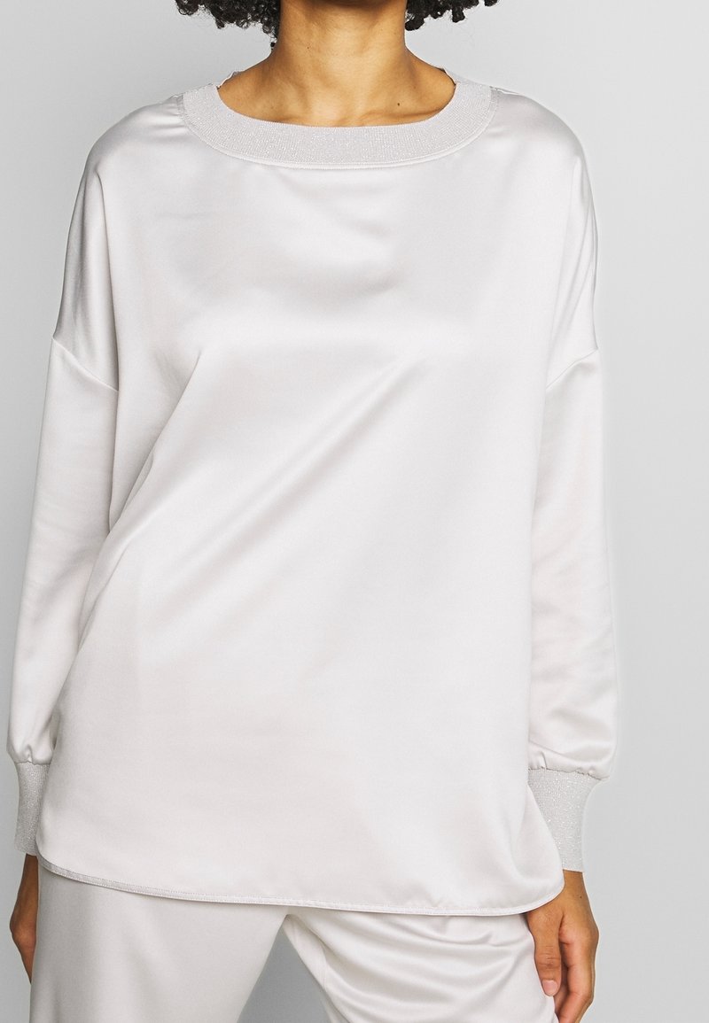 Mujer vistiendo una blusa larga de satén blanca y suelta con puños y escote acanalados brillantes, combinada con pantalones de satén blancos a juego.