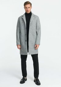 Hallhall, tekstuuriga villane mantel kraega, kahe musta nupuga ja küljetaskutega, kombineeritud musta kõrge kaelusega särkide ja mustade pükstega.