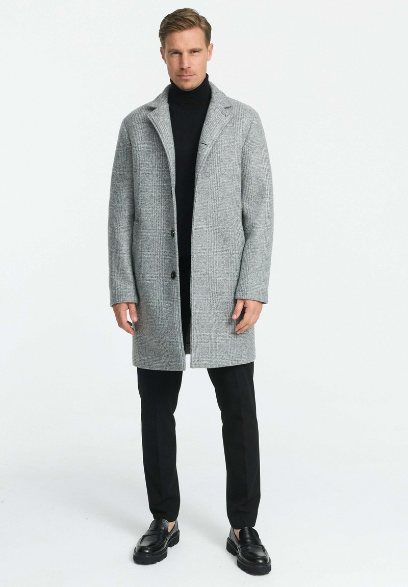 Hallhall, tekstuuriga villane mantel kraega, kahe musta nupuga ja küljetaskutega, kombineeritud musta kõrge kaelusega särkide ja mustade pükstega.