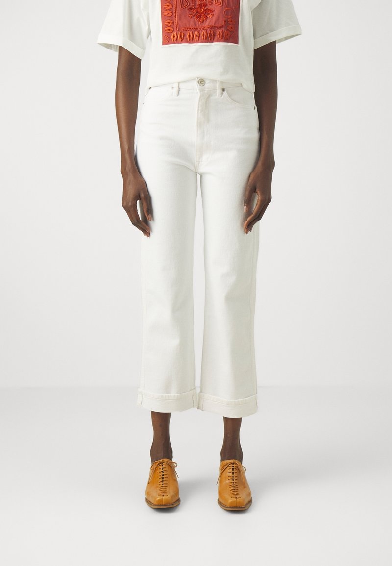 Personne portant un jean blanc taille haute coupé, un t-shirt blanc avec une broderie orange et des chaussures en cuir marron clair à lacets, debout sur un sol blanc.