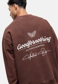 Felpa marrone con grafica di farfalla e scritta "Goodfornothing" sul retro, con dettagli bianchi e una vestibilità rilassata.