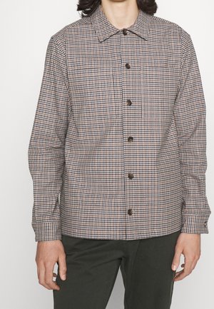 Man draagt een shirt met lange mouwen en knopen met een geruit patroon in bruin, wit en zwart, gecombineerd met donkergroene broek.