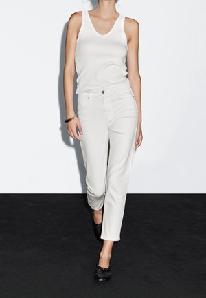 Femme portant un haut blanc sans manches, un pantalon blanc taille haute, des chaussures noires et un bracelet en or, debout contre un mur blanc et un sol noir.